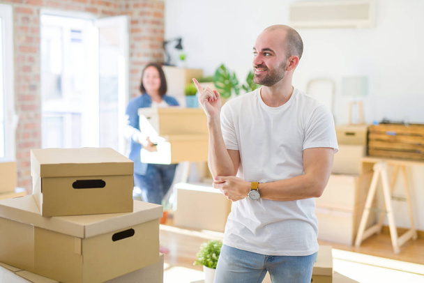 Young couple arround cardboard boxes moving to a new house, bald man standing at home with a big smile on face, pointing with hand and finger to the side looking at the camera. - 写真・画像