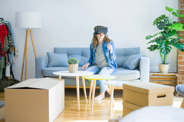 Young blonde woman wearing virtual reality glasses playing a simulation game sitting on the sofa around cardboard boxes moving to a new house - Фото, изображение