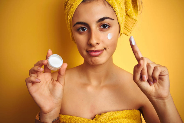 Young woman wearing a shower towel using face cream over yellow isolated background with a confident expression on smart face thinking serious - Photo, Image