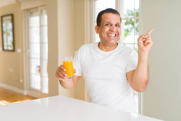 Μέση ηλικία άνθρωπος πίνοντας ένα ποτήρι πορτοκάλι χυμό στο σπίτι πολύ χαρούμενος, δείχνοντας με το χέρι και δάχτυλο προς την πλευρά - Φωτογραφία, εικόνα