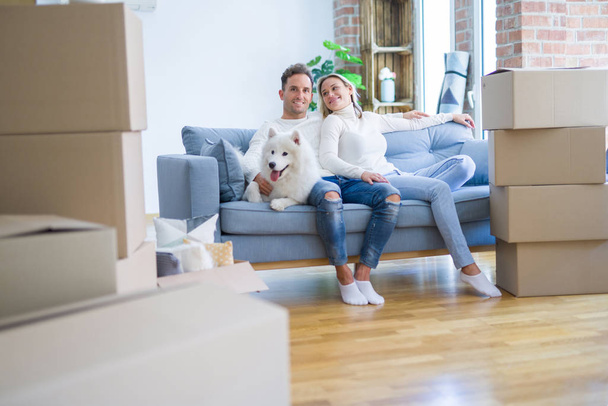 Joven hermosa pareja con perro sentado en el sofá en el nuevo hogar alrededor de cajas de cartón
 - Foto, imagen