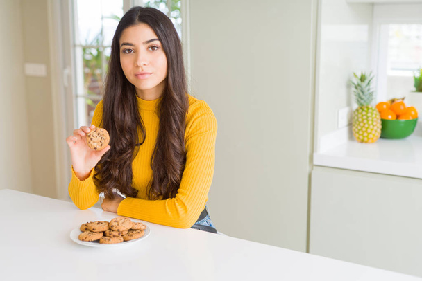 Mujer joven comiendo galletas chips de chocolate en casa con una expresión de confianza en la cara inteligente pensando en serio
 - Foto, Imagen