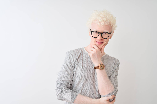 Jonge albino blonde man draagt gestreepte t-shirt en glazen over geïsoleerde witte achtergrond kijken zelfverzekerd naar de camera glimlachend met gekruiste armen en hand verhoogd op de kin. Positief denken. - Foto, afbeelding
