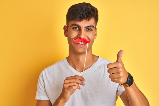 Jeune homme indien tenant une moustache fanny debout sur un fond jaune isolé heureux avec un grand sourire faisant signe ok, pouce levé avec les doigts, excellent signe
 - Photo, image