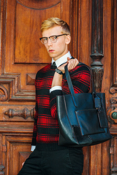 Young blonde American male college student wearing patterned red, black knit sweater, glasses, holding leather bag on shoulder, standing by brown vintage wooden office door, looking at you - Photo, Image