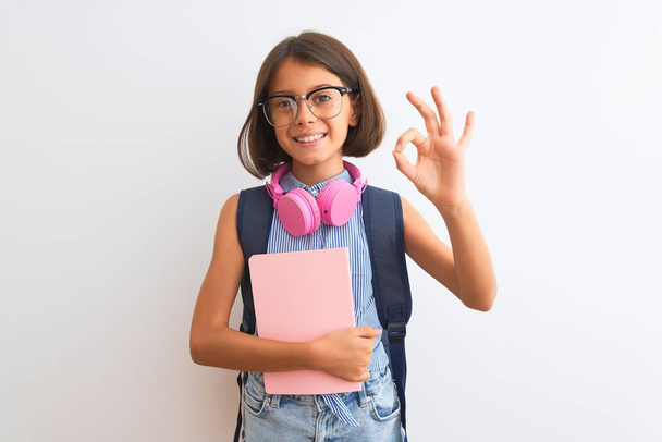 Student dziecko dziewczyna noszenie plecak okulary książki słuchawki na białym tle robi OK znak z palcami, doskonały symbol - Zdjęcie, obraz