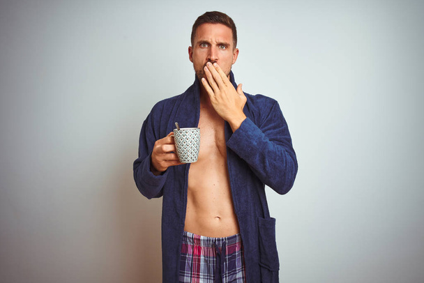 Un hombre sin camisa que lleva un pijama cómodo y una túnica bebiendo una taza de café cubre la boca con la mano conmocionada con vergüenza por error, expresión de miedo, asustado en silencio, concepto secreto
 - Foto, imagen