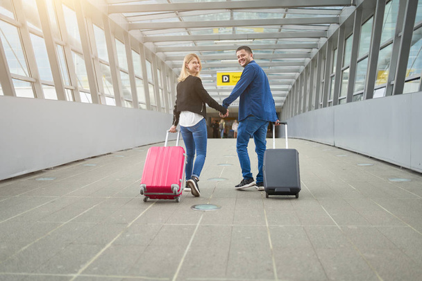 Фото сзади мужчины и женщины, смотрящих в камеру, гуляющих с чемоданами по проходу в аэропорту
 - Фото, изображение