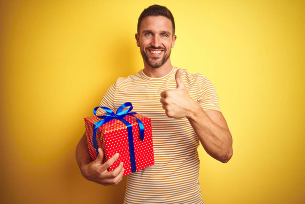 Νέος άνθρωπος κρατώντας ένα δώρο γενεθλίων πάνω από κίτρινο απομονωμένο φόντο χαρούμενο με μεγάλο χαμόγελο κάνει OK πινακίδα, αντίχειρας με τα δάχτυλα, εξαιρετική πινακίδα - Φωτογραφία, εικόνα
