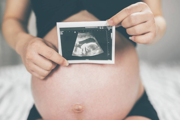 Εγκυμοσύνης. ένα πορτραίτο ενός ζευγαριού μικρών υποδημάτων για το αγέννητο κοντά του με μια έγκυο γυναίκα που αγκαλιάζει την κοιλιά της με αγάπη, ζεστασιά στο σπίτι στο υπνοδωμάτιο το φόντο, αντίγραφο χώρου - Φωτογραφία, εικόνα