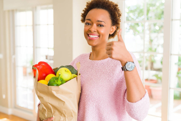 Young african american woman holding paper bag full of fresh groceries happy with big smile doing ok sign, thumb up with fingers, excellent sign - Foto, afbeelding