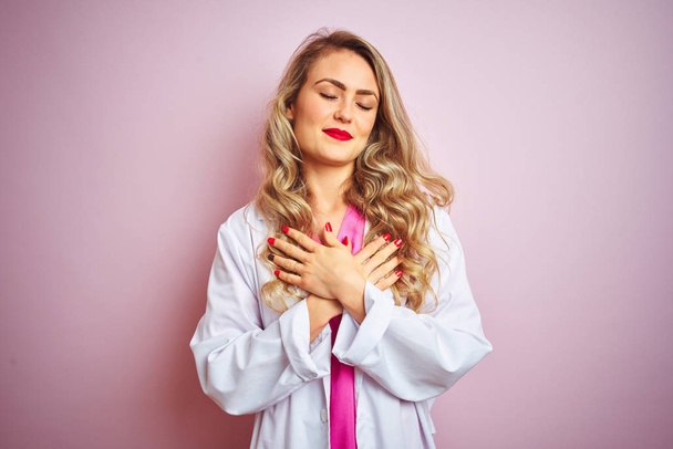 Young beautiful doctor woman standing over pink isolated background smiling with hands on chest with closed eyes and grateful gesture on face. Health concept. - Photo, Image