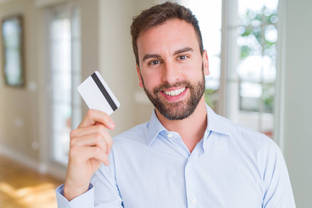 Yakışıklı bir iş adamı elinde gülen bir yüzle ayakta duran ve gülümseyen dişlerini gösteren bir kredi kartı tutuyor. - Fotoğraf, Görsel