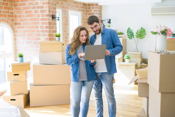 Young couple using computer laptop standing on a room around cardboard boxes, happy for moving to a new apartment - Photo, Image