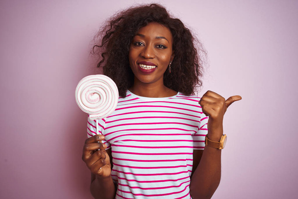 Joven mujer afroamericana comiendo dulces de pie sobre un fondo rosa aislado señalando y mostrando con el pulgar hacia el lado con la cara feliz sonriendo
 - Foto, imagen