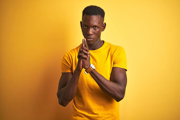 Young african american man wearing casual t-shirt standing over isolated yellow background Holding symbolic gun with hand gesture, playing killing shooting weapons, angry face - Фото, изображение
