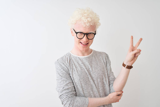 Młody Albino blond człowiek ubrany w paski t-shirt i okulary na białym tle, uśmiechając się szczęśliwy twarz mżąc w aparacie robi znak zwycięstwa. Numer dwa. - Zdjęcie, obraz