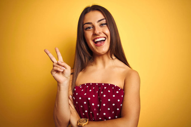 Young beautiful woman wearing red t-shirt standing over isolated yellow background smiling with happy face winking at the camera doing victory sign. Number two. - 写真・画像