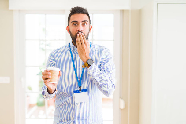 Handsome hispanic man wearing id card and drinking a cup of coffee cover mouth with hand shocked with shame for mistake, expression of fear, scared in silence, secret concept - Photo, image