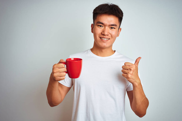 Asian chinese man holding red cup of coffee standing over isolated white background happy with big smile doing ok sign, thumb up with fingers, excellent sign - Fotografie, Obrázek