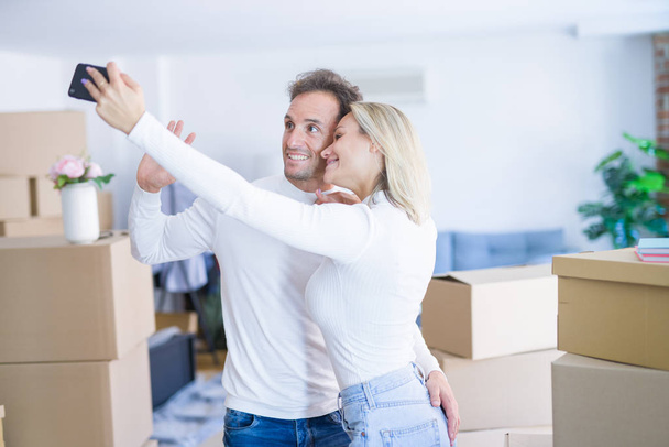 Νεαρό όμορφο ζευγάρι στέκεται χρησιμοποιώντας το smartphone για να πάρει selfie στο νέο σπίτι γύρω από χαρτόνι κουτιά - Φωτογραφία, εικόνα