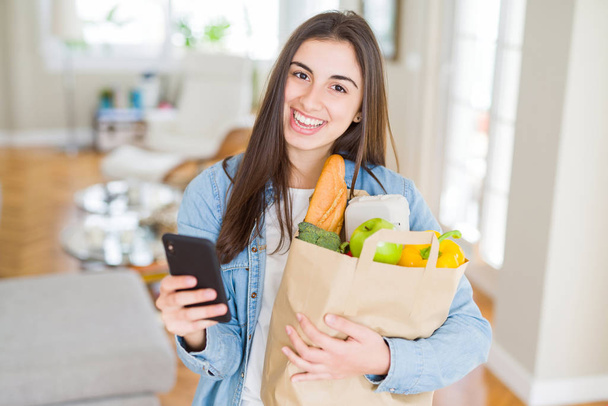 Young woman holding a paper bag full of fresh groceries and usin - Foto, imagen