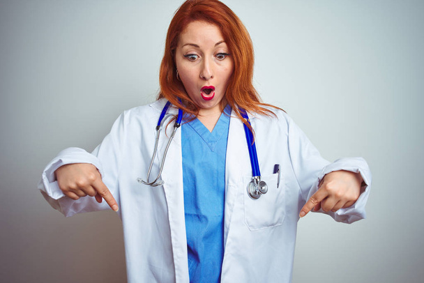 Молодая рыжая женщина-врач, использующая стетоскоп на белом изолированном фоне, указывает пальцами вниз, показывая рекламу, удивленное лицо и открытый рот
 - Фото, изображение