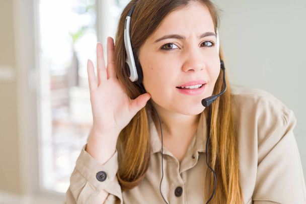 Mooie jonge operator vrouw het dragen van headset op het kantoor glimlachend met de hand over het luisteren van een gehoor tot geruis of roddels. Doofheid concept. - Foto, afbeelding