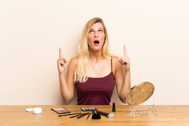 Mujer rubia joven con cosmética en una mesa señalando con el dedo índice una gran idea
 - Foto, Imagen