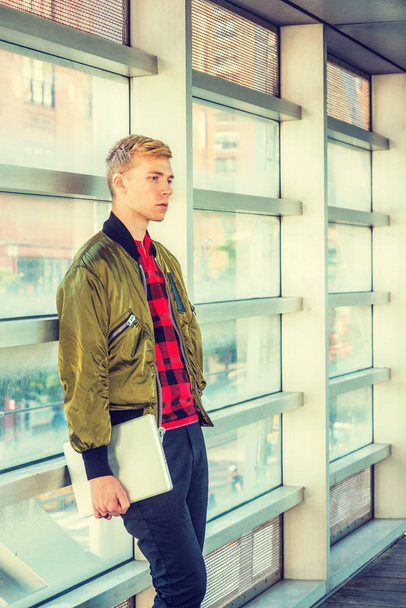 Young blonde American college student wearing green jacket unzipped, red patterned shirt, holding laptop computer, standing on walkway against glass wall on campus in New York, thinking - Фото, изображение