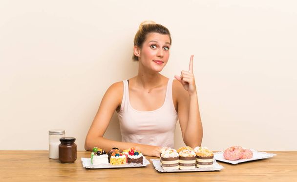 junge Frau mit vielen verschiedenen Mini-Kuchen in einem Tisch, der mit dem Zeigefinger zeigt eine großartige Idee - Foto, Bild