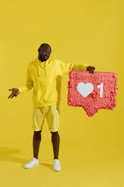 Like. Disappointed man holding heart icon pinata on yellow  - Φωτογραφία, εικόνα