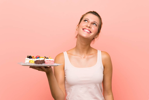 Young blonde woman holding lots of different mini cakes looking up while smiling - Фото, зображення