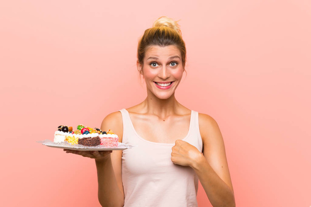 Young blonde woman holding lots of different mini cakes with surprise facial expression - Foto, Imagen