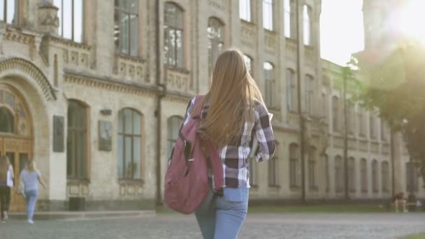 Opiskelijatyttö kävelee yliopistorakennukseen. Iloinen ja hymyilevä, katsoo kameraan ja kävelee eteenpäin. Näkymä takaa. Hidastus
. - Materiaali, video