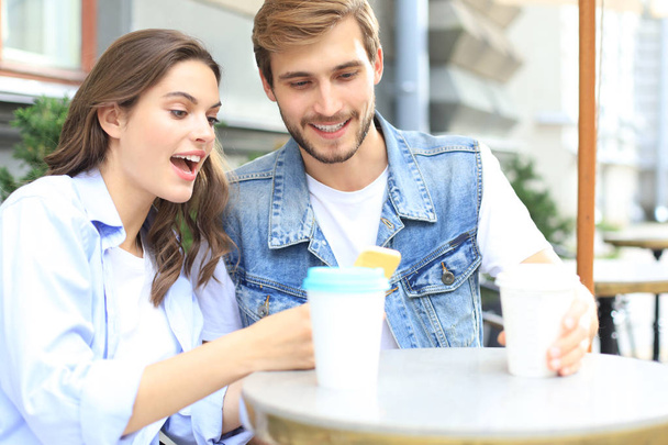 Attractive young couple in love drinking coffee while sitting at the cafe table outdoors, using mobile phone. - Foto, Imagem