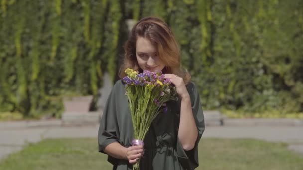 Portrait of playful cute girl sniffing wild flowers looking at camera while standing outdoors. Concept of summertime, rest day. Real people series. - Imágenes, Vídeo