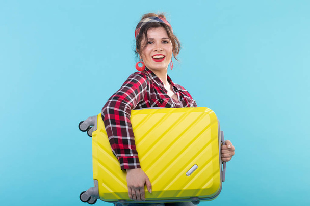 Joven mujer sonriente positiva con una camisa a cuadros que sostiene una maleta amarilla posando sobre un fondo azul. Concepto de turismo y viajes
 - Foto, imagen