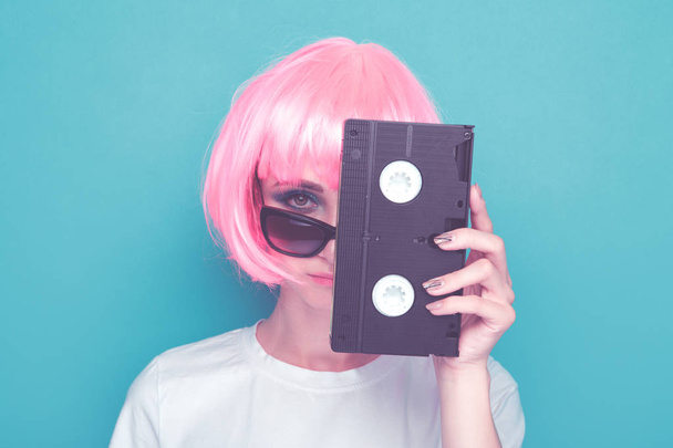 Retro dívka v brýlích a růžové paruce držící černou kazetu na modrém pozadí. - Fotografie, Obrázek