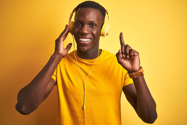 Izole sarı arka plan üzerinde kulaklık kullanarak müzik dinleyen Afrikalı Amerikalı adam mutlu yüz, bir numaralı ile bir fikir ya da soru işaret parmağı ile şaşırttı - Fotoğraf, Görsel