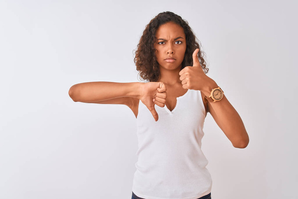 Mladá brazilská žena, která nosí neformální tričko stojící nad izolovaným bílým pozadím dělá palce nahoru a dolů, nesouhlas a výraz souhlasu. Šílený konflikt - Fotografie, Obrázek