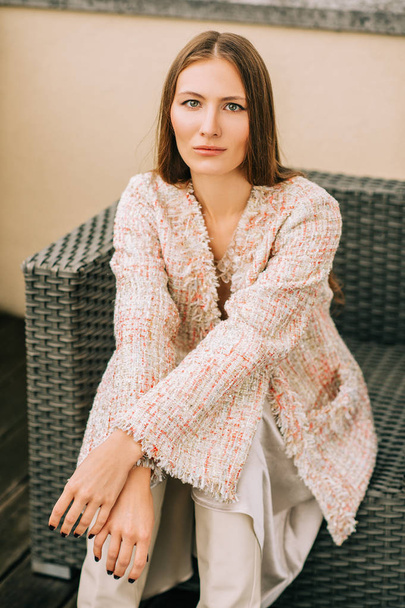 Outdoor fashion portrait of young woman wearing stylish peach colour jacket and silk skirt, sitting on the chair - Foto, afbeelding