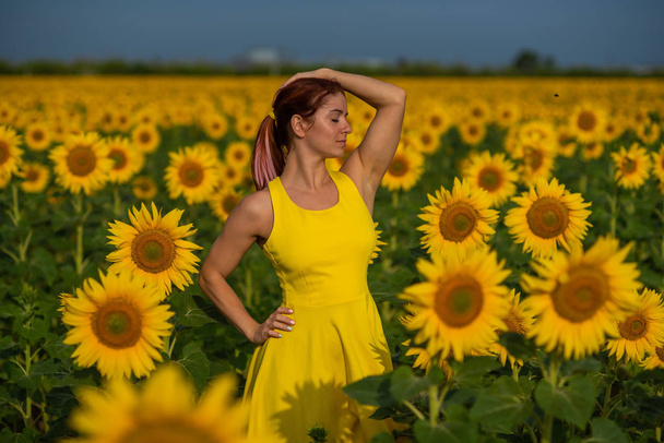 A red-haired woman in a yellow dress is standing in a field of sunflowers. Beautiful girl in a skirt sun enjoys a cloudless day in the countryside. Pink locks of hair. - Фото, изображение