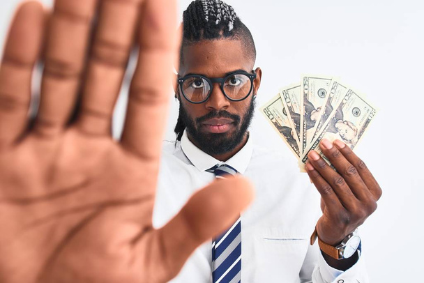 African american businessman with braids holding dollars over isolated white background with open hand doing stop sign with serious and confident expression, defense gesture - Foto, afbeelding