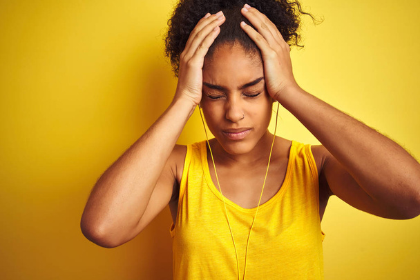 Афроамериканець жінка слухає музику за допомогою навушники на ізольований жовтий фон страждає від головного болю відчайдушним і підкреслив, тому що біль і мігрень. Руки на голові. - Фото, зображення