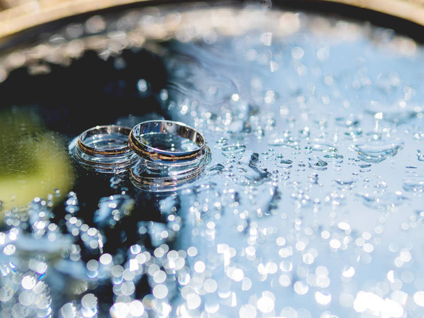 Dvojice svatebních kroužků na mokrém zrcadlovém povrchu s odrazem. Zavřít fotografii s tradičními šperky nevěstou a ženichem. - Fotografie, Obrázek