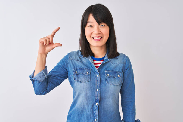 Genç güzel Çinli kadın izole beyaz arka plan üzerinde denim gömlek giyen gülümseyen ve emin el parmakları görünümlü ve kamera ile küçük boyutlu işareti yapıyor gesturing. Ölçü kavramı. - Fotoğraf, Görsel