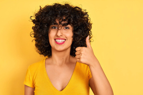 Young arab woman with curly hair wearing t-shirt standing over isolated yellow background happy with big smile doing ok sign, thumb up with fingers, excellent sign - Foto, afbeelding