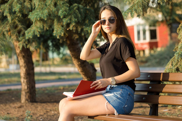 Ένα νεαρό κορίτσι κάθεται σε ένα παγκάκι σε ένα πάρκο και κάνει σημειώσεις. Ντυμένος με ελεύθερο στυλ. Επιχείρηση γυναίκα μιλάει στο τηλέφωνο με τους πελάτες. - Φωτογραφία, εικόνα