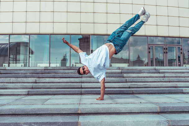 athlete guy dancer in white t-shirt, jeans, jumped on one arm, in summer in city, in sunglasses, stairs background, active hip hop, youth lifestyle, free space for text, acrobatic stunt break dance - Foto, Bild